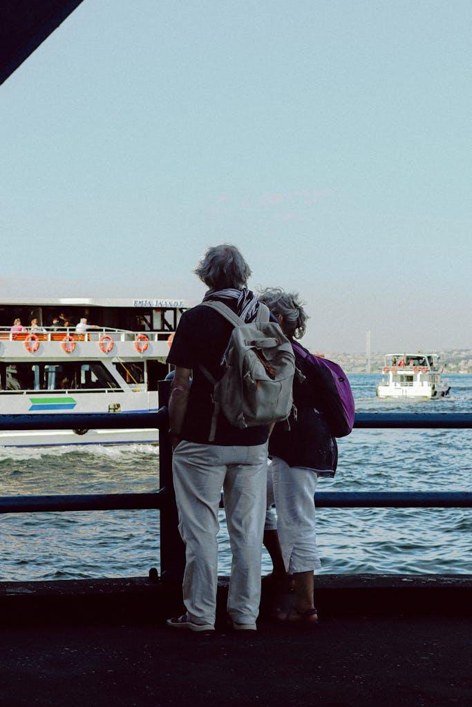Elderly couple stands by the waterfront watching boats on a sunny day.